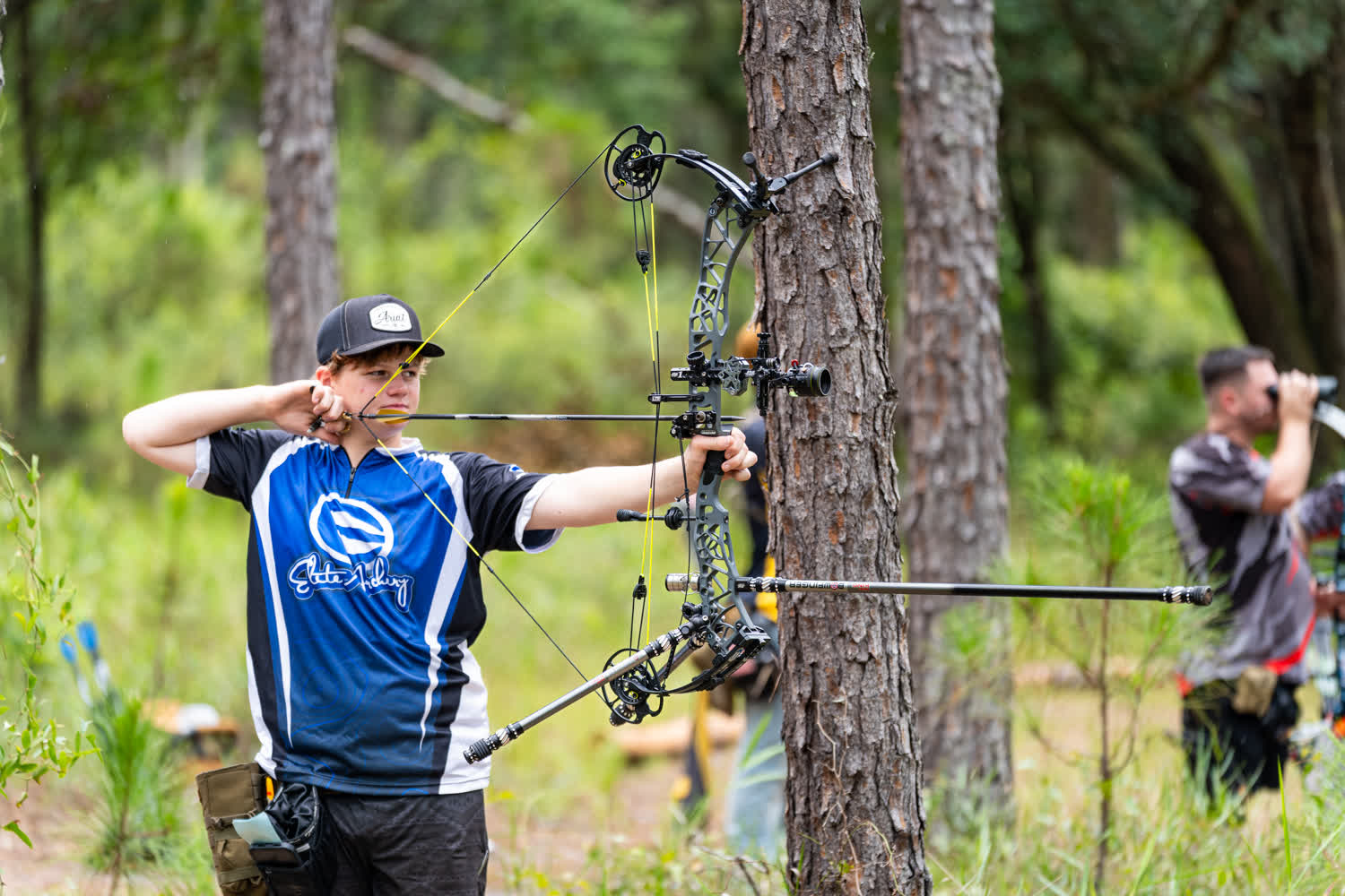 Young archer in a blue and white jersey and dark cap at full draw with a compound bow among pine trees on a wooded course, with a man looking through binoculars in the background.