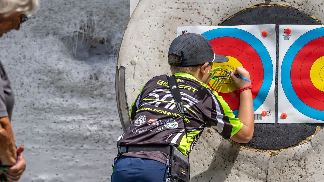 Young archer in a Sarasota Archery Academy jersey and cap, seen from behind, marking arrow holes on a paper target face while an adult stands at the left edge.
