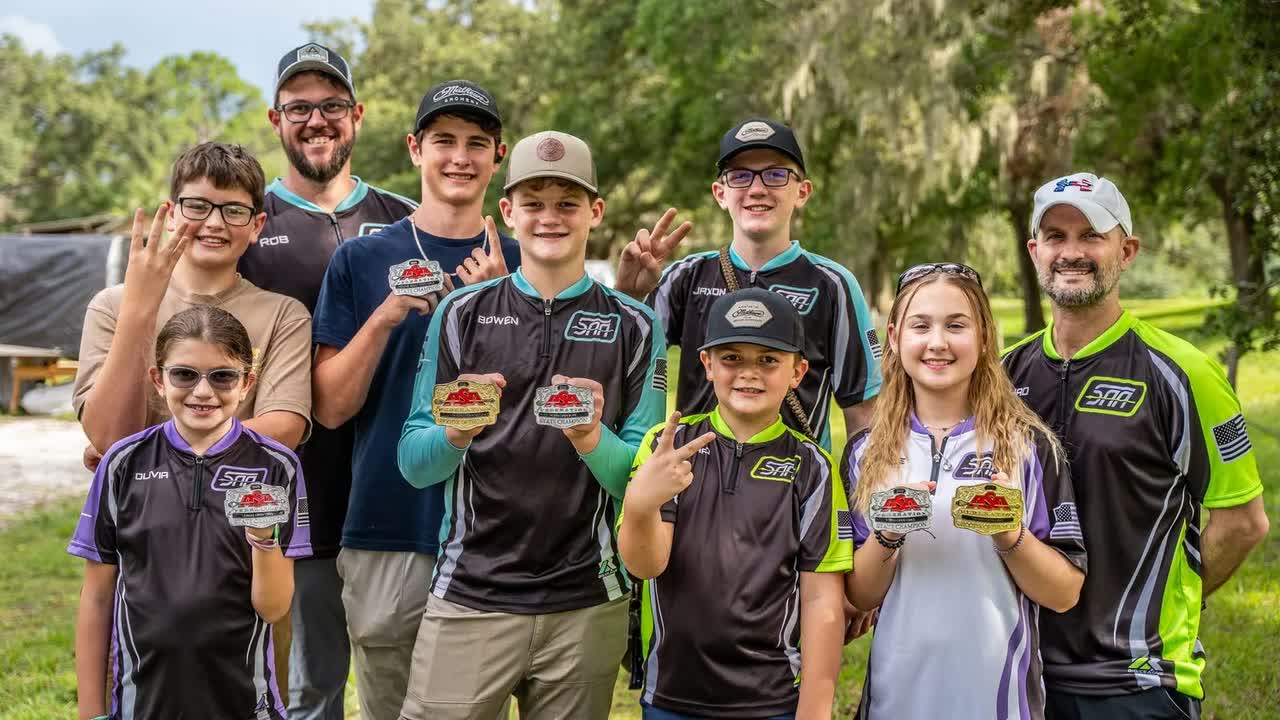 Youth archers in Sarasota Archery Academy jerseys holding up medals outdoors, with two adults in team jerseys at the back and right.