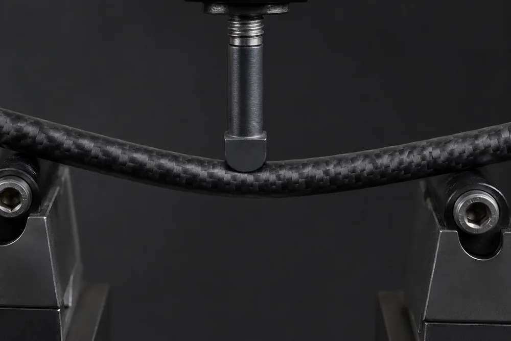 Close-up of a carbon arrow shaft resting on two supports with a weight pressing its centre in a spine-testing fixture, showing the shaft flexing.