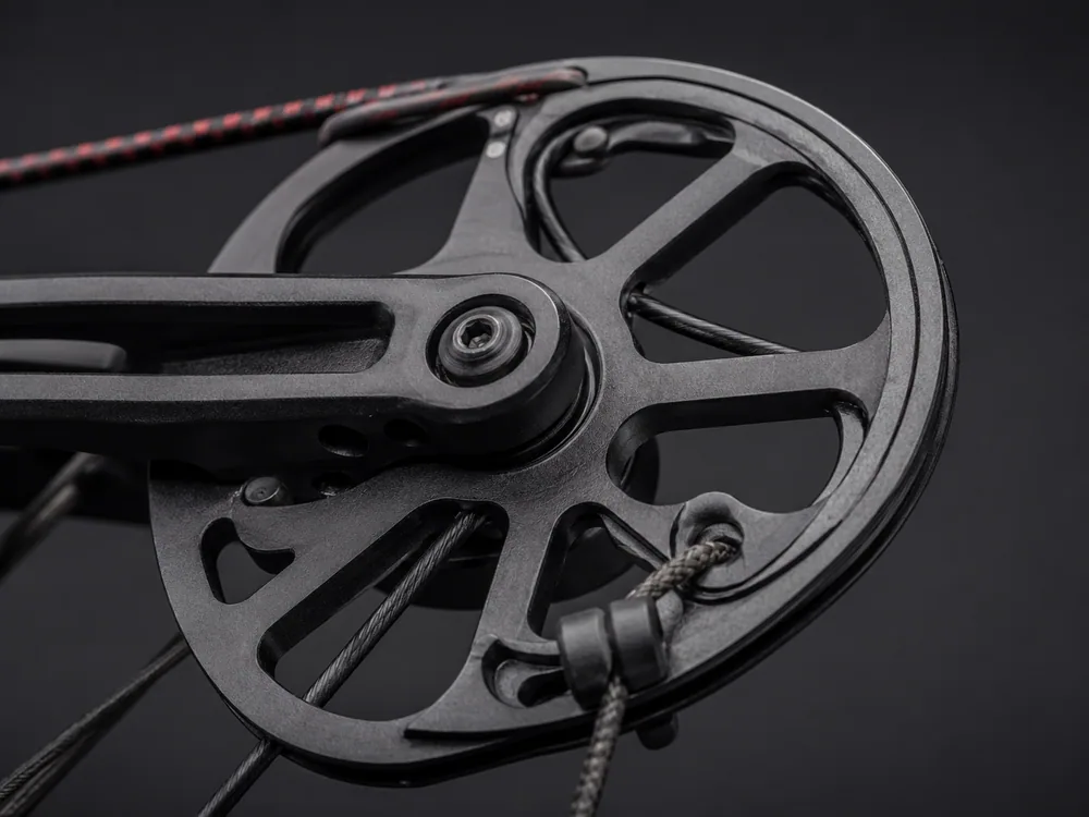 Close-up of a black machined compound bow cam at the end of a limb, with the bowstring and cable routed around its grooves, on a dark background.