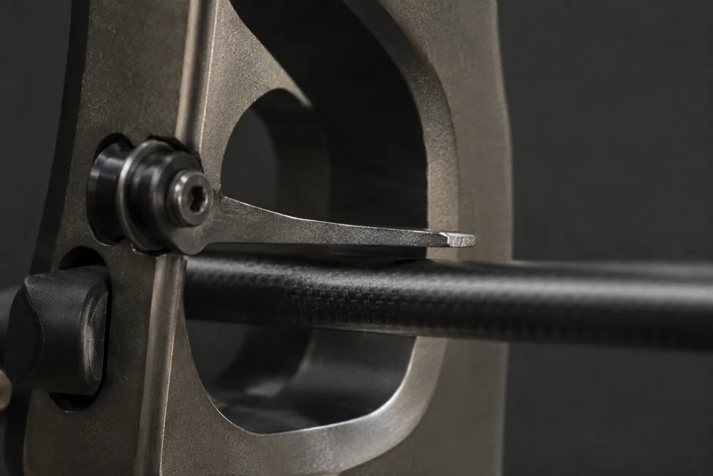 Close-up of a thin spring-steel clicker blade mounted on a recurve bow riser window with an arrow shaft beneath it.