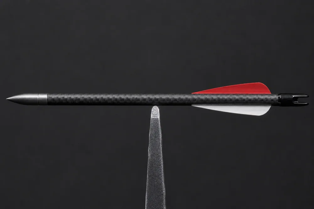 A finished carbon arrow balanced at its balance point on a narrow fulcrum edge — the measurement FOC is calculated from.